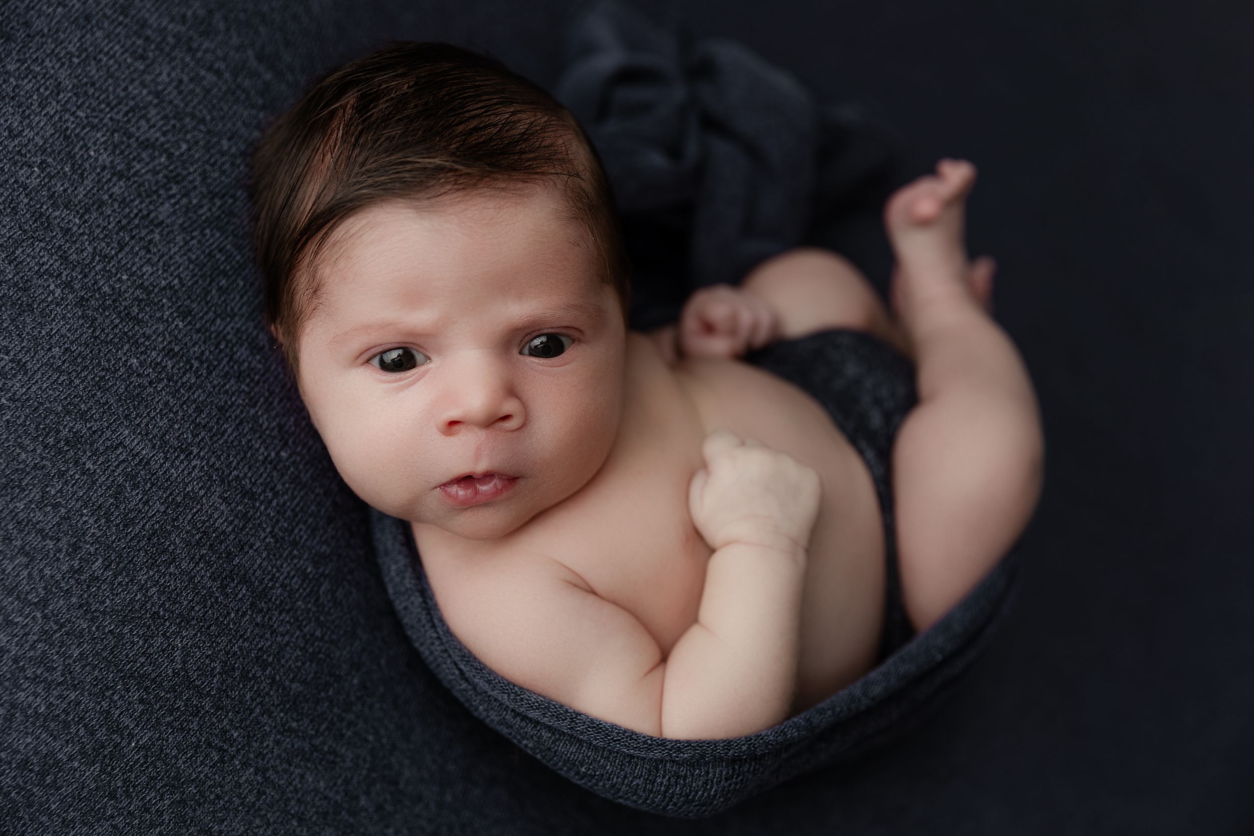 A newborn baby lies on dark fabric in a circular basket during a professional portrait session.