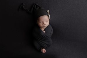 A newborn baby peacefully sleeping on a black blanket while wrapped in dark cloth and wearing a knit cap.