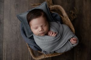 Peaceful sleeping newborn swaddled in soft gray fabric resting in rustic wooden basket.