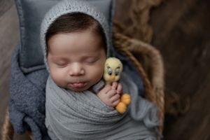 Peaceful sleeping baby wrapped in grey swaddle with yellow toy duck nestled close, against dark background.