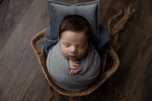 Sleeping newborn baby wrapped in gray swaddle lying in woven basket with pillow on wooden floor.