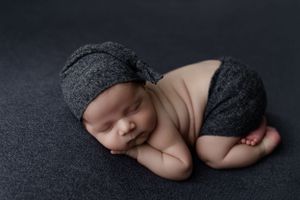Sleeping newborn baby wearing gray knit hat and shorts lying on dark backdrop during studio photoshoot.