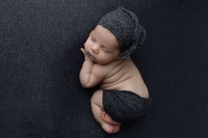 Sleeping newborn baby wearing dark knit bonnet and wrap curled up on black backdrop during professional photo session.