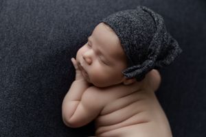Sleeping newborn baby wearing gray knit hat lying on dark backdrop during professional photo session.