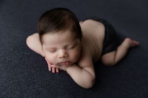 Peaceful sleeping newborn baby wearing dark bottoms lying on charcoal grey blanket during professional photo session.