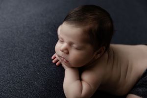 Peaceful sleeping newborn baby rests their head on folded hands against dark textured background.