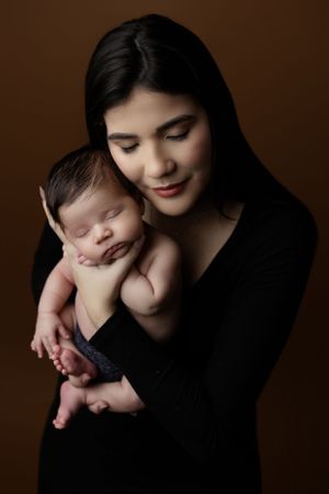 A tender portrait of mother in black clothing holding sleeping newborn baby against brown backdrop.