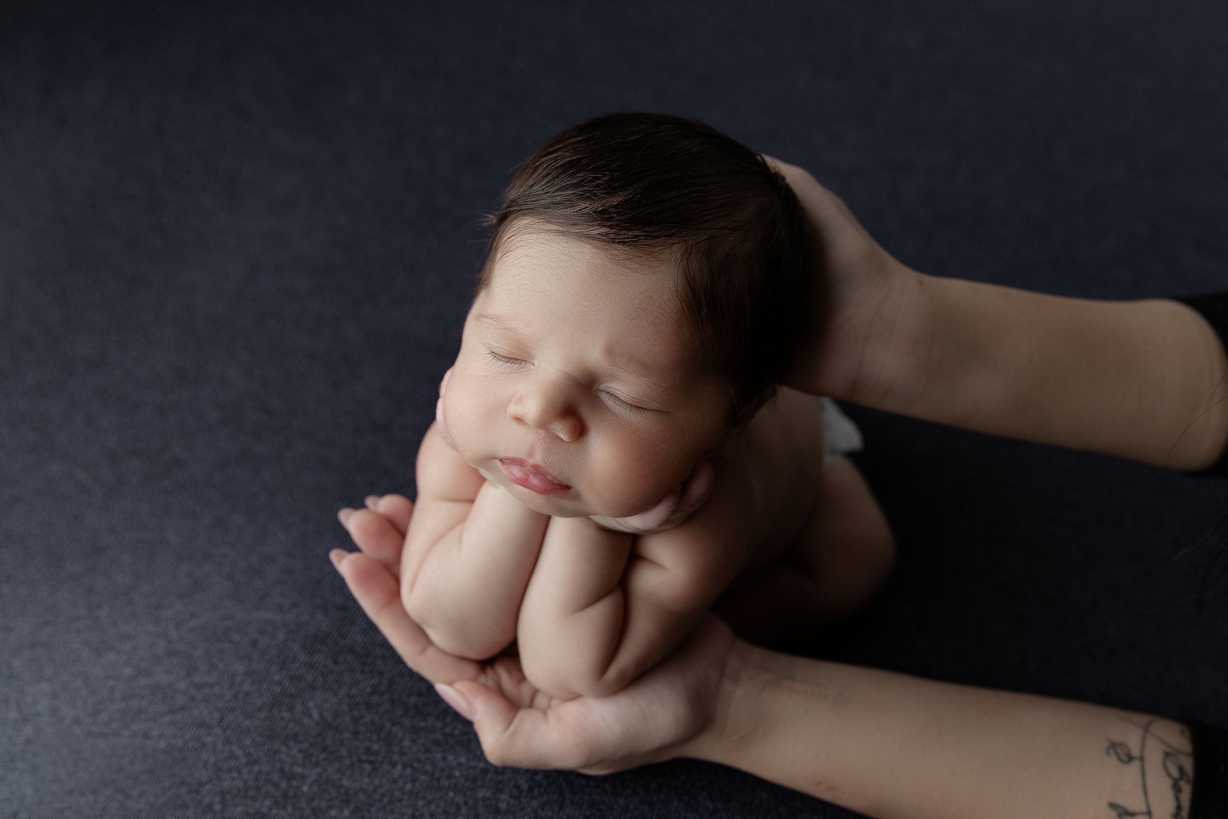 Newborn baby sleeping peacefully while being gently cradled in adult hands against dark background.