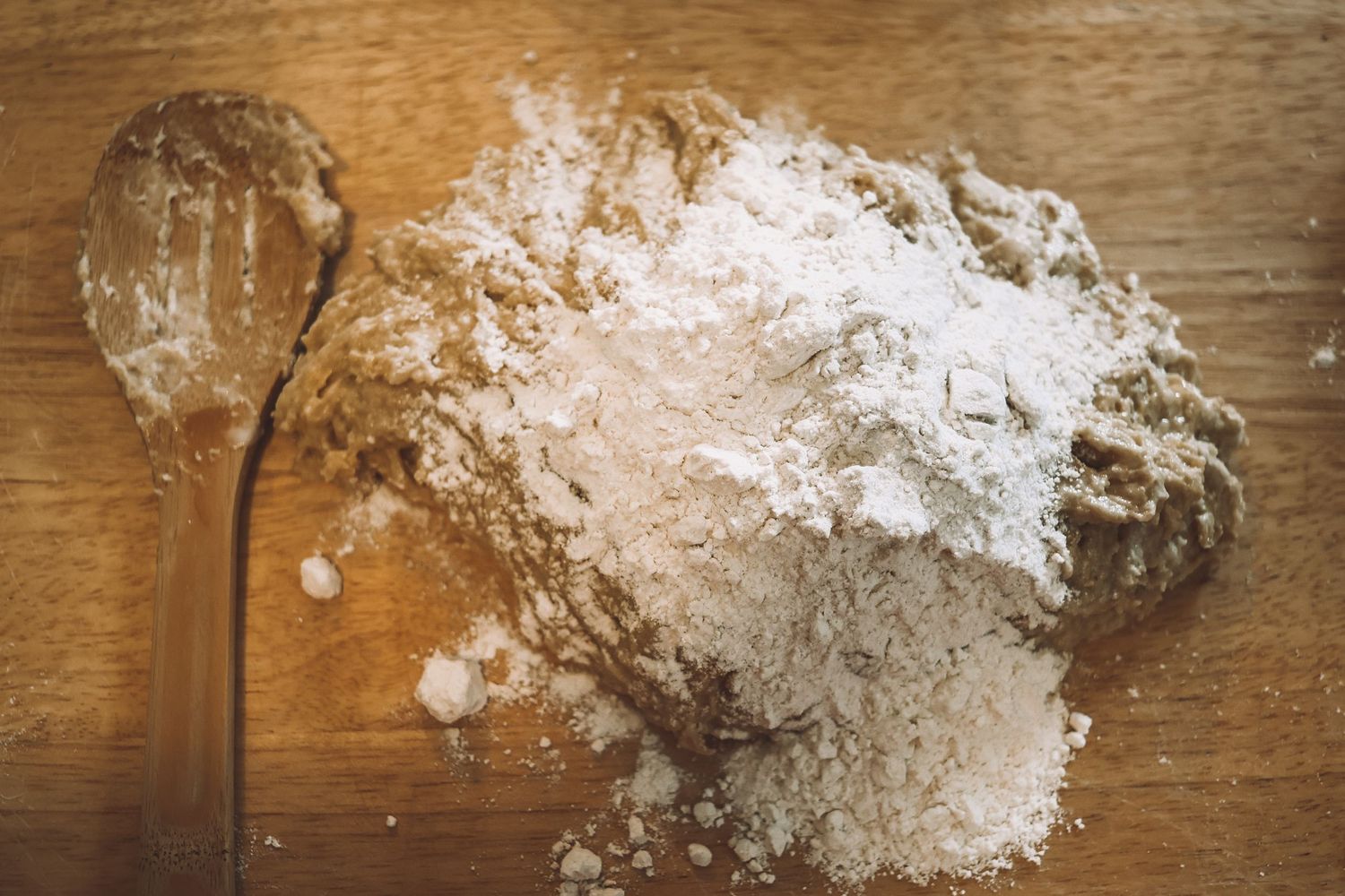White flour scattered on a wooden cutting board with a measuring spoon beside it.