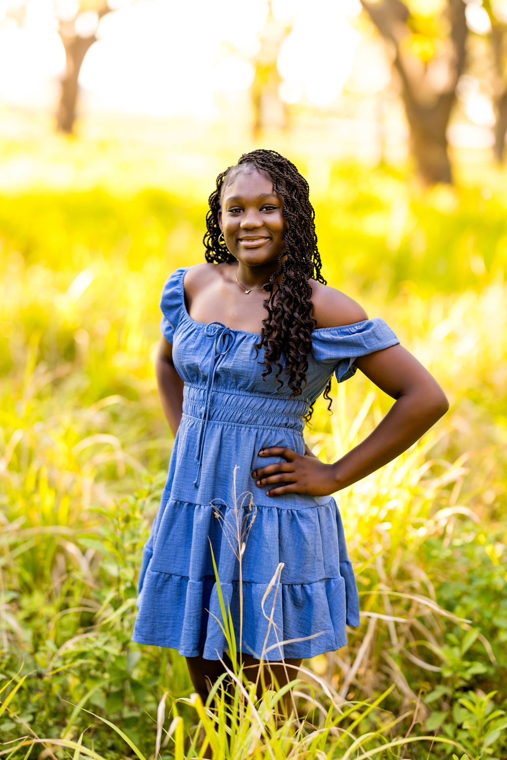 A person poses in a blue off-shoulder dress against a sunlit field of tall grass.