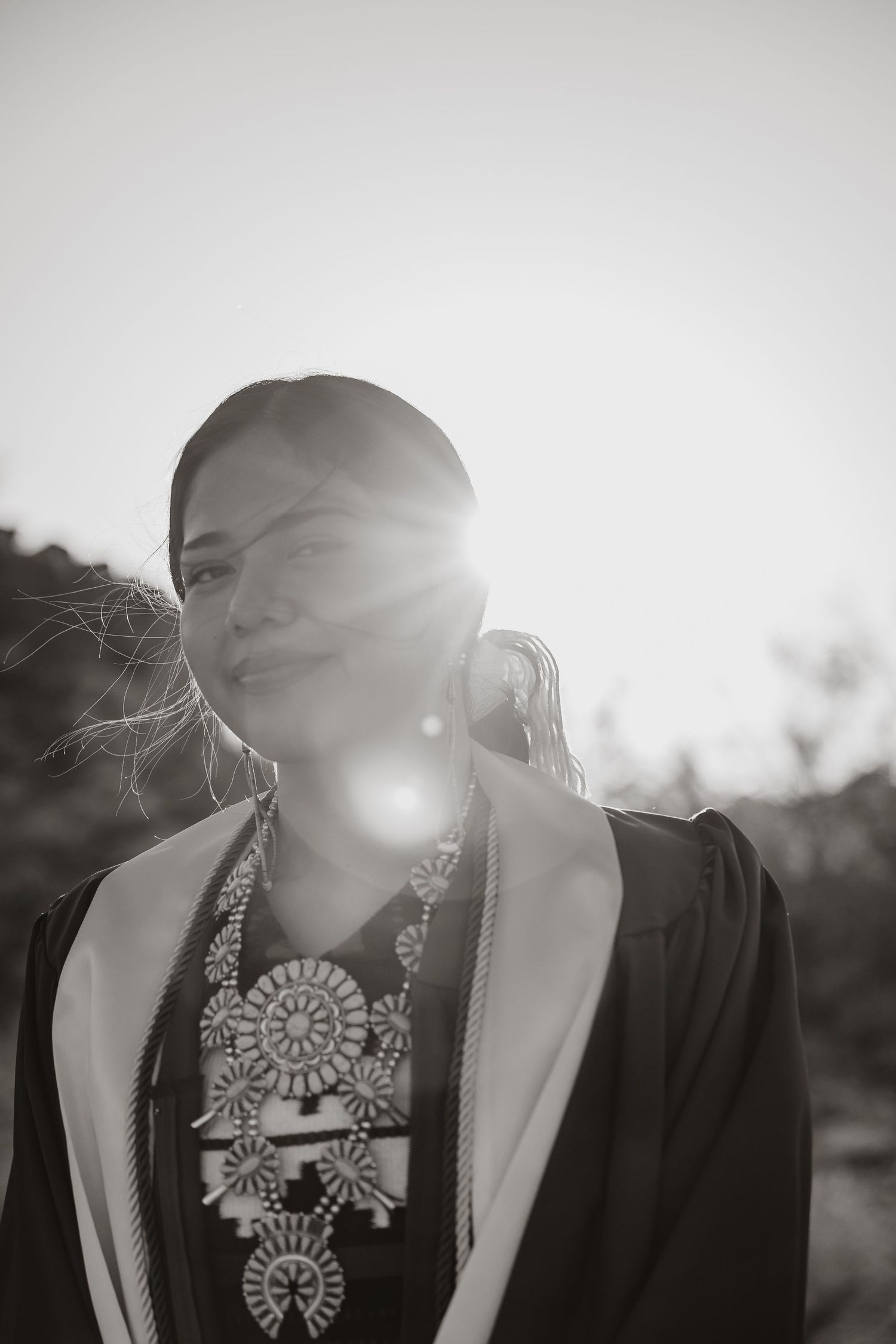 Black and white portrait of a young woman in graduation gown and Native American jewelry, backlit by sunlight.