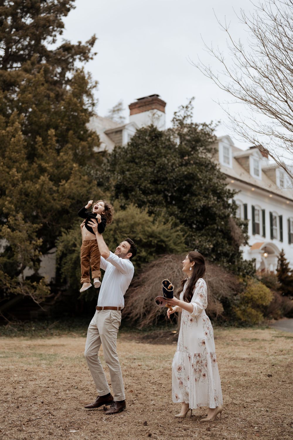 A couple plays with their child in front of a white colonial-style house.