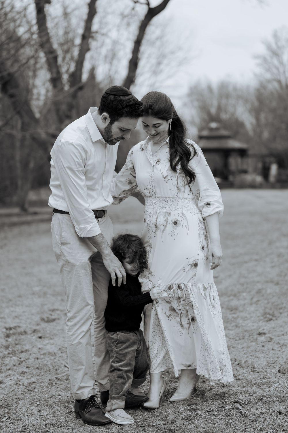A couple in white outfits poses outdoors with a small child in a casual family photoshoot setting.