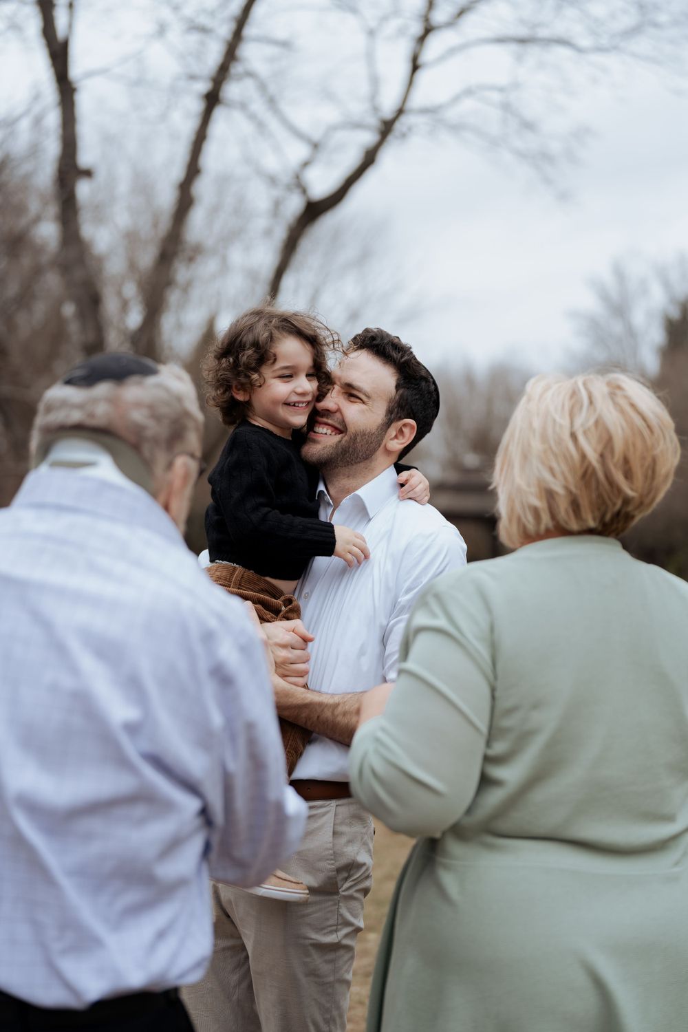 Family members share an intimate moment outdoors while an older couple looks on from the side.
