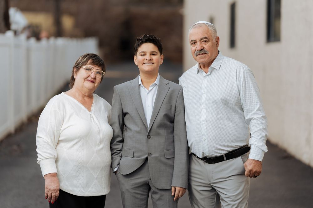 Three generations pose together against a white wall in casual formal attire.