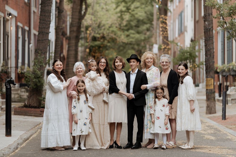 Group of people wearing formal white and black attire pose together on a charming brick-lined city street in springtime.