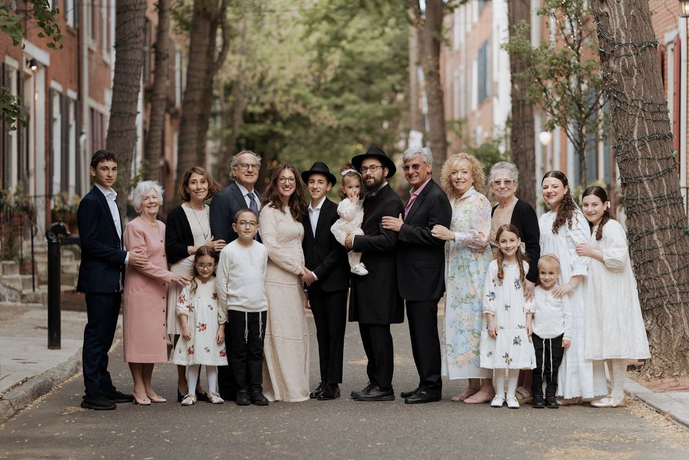 Large group portrait of well-dressed people standing together on a tree-lined brick street.