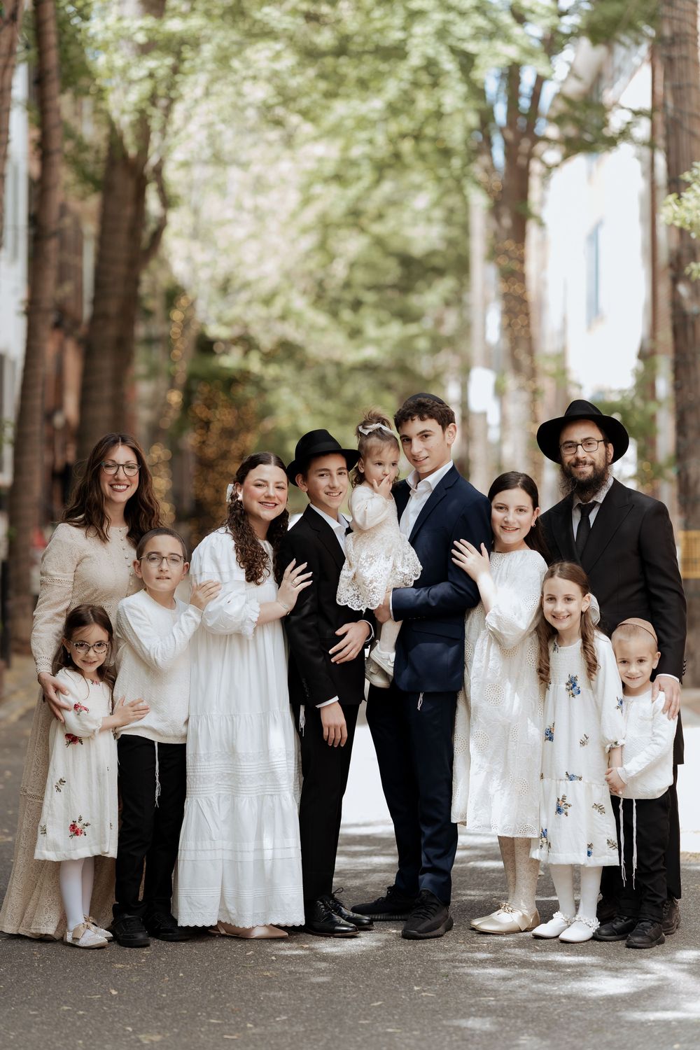 A group in formal attire stands together on a tree-lined path, wearing black and white clothing in a coordinated style.