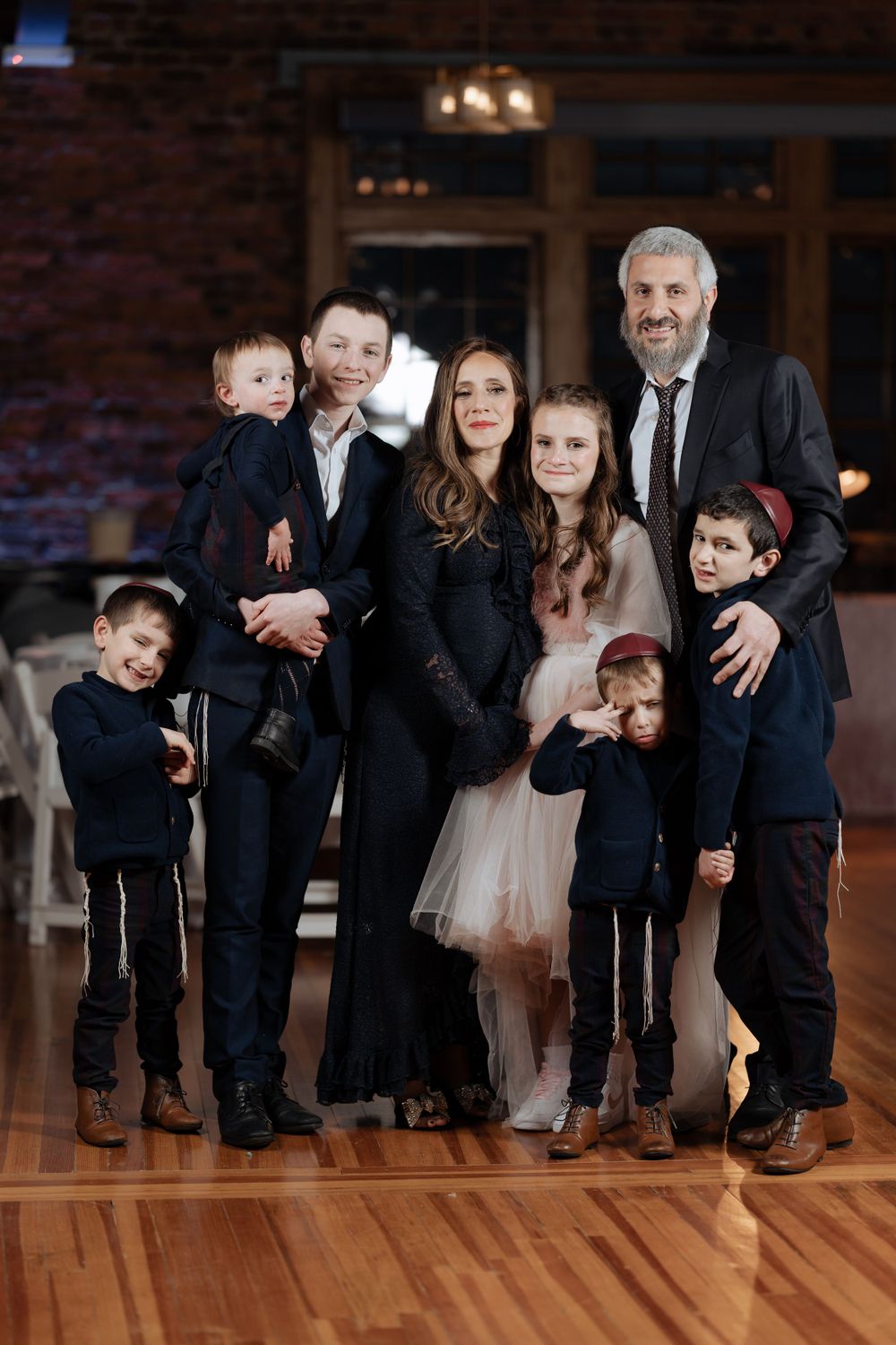 A formal indoor family portrait with everyone dressed in dark attire against a brick wall background.