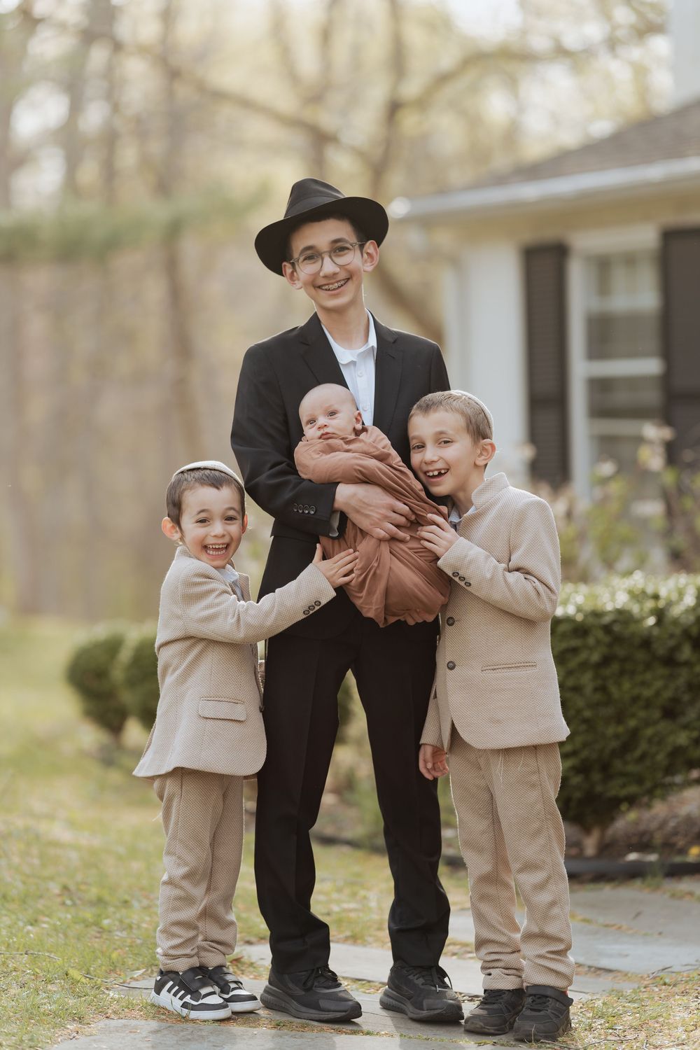 A figure in a black suit and hat stands outdoors with three children in beige suits in front of a house.