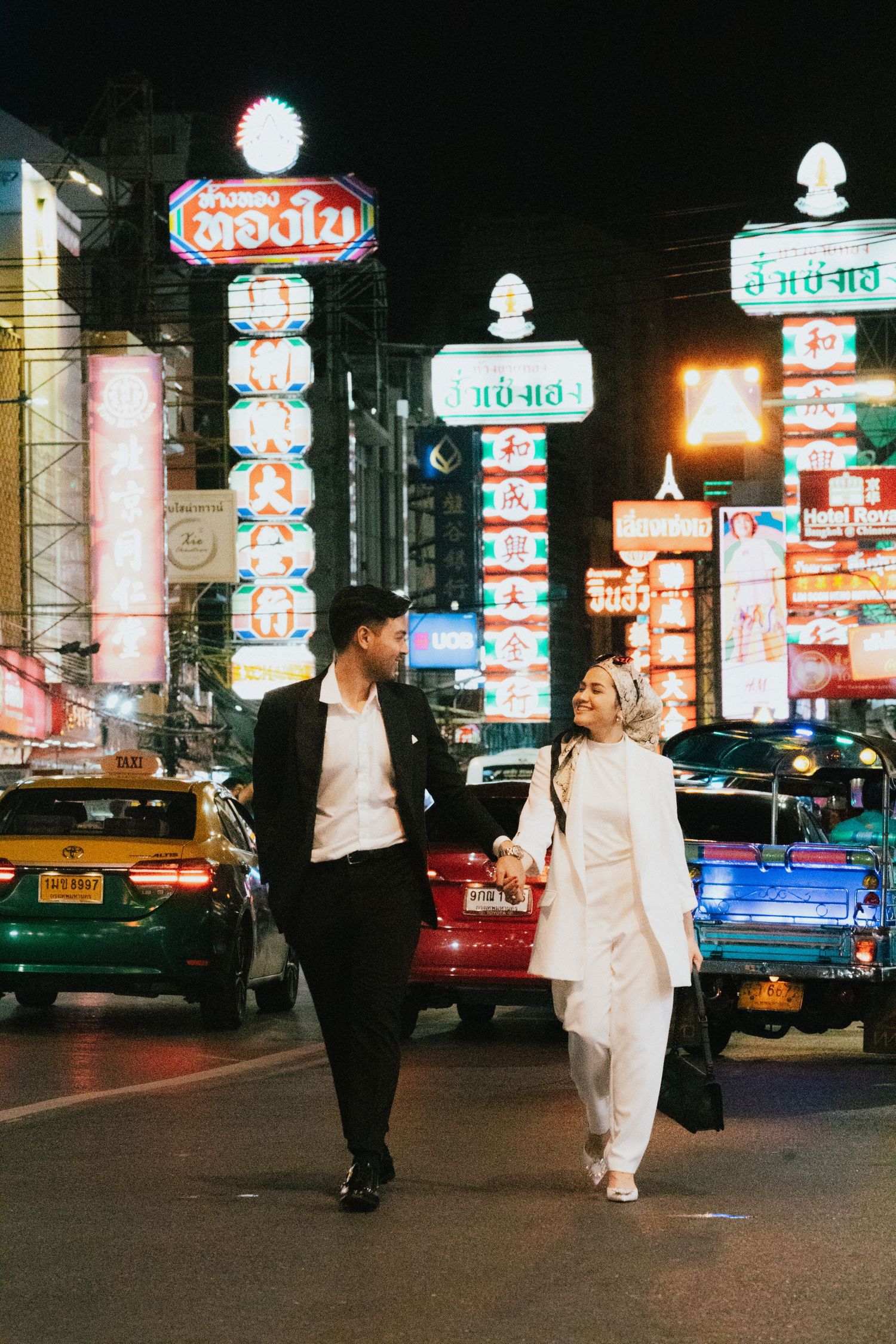 Couple holding hands walking through Bangkok Chinatown at night with colorful neon signs glowing behind them.