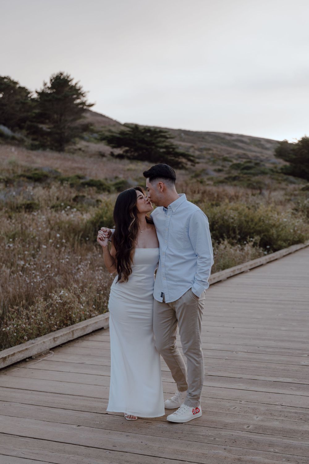 A couple sharing a romantic moment on a wooden boardwalk against a backdrop of rolling hills at sunset.