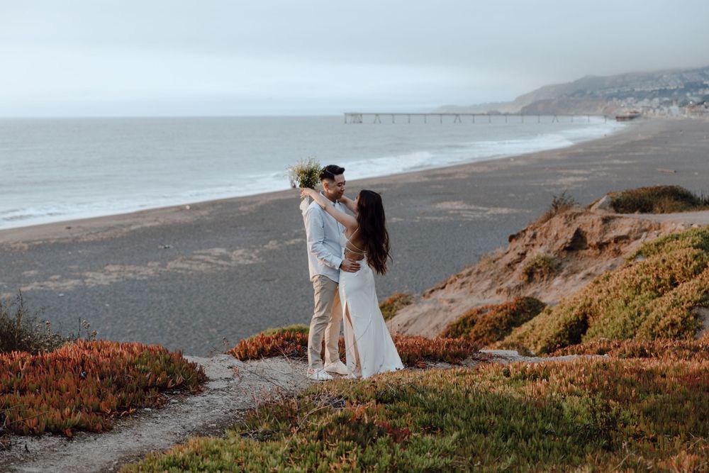 A couple embraces on a coastal clifftop overlooking a misty beach at sunset with orange wildflowers in the foreground.