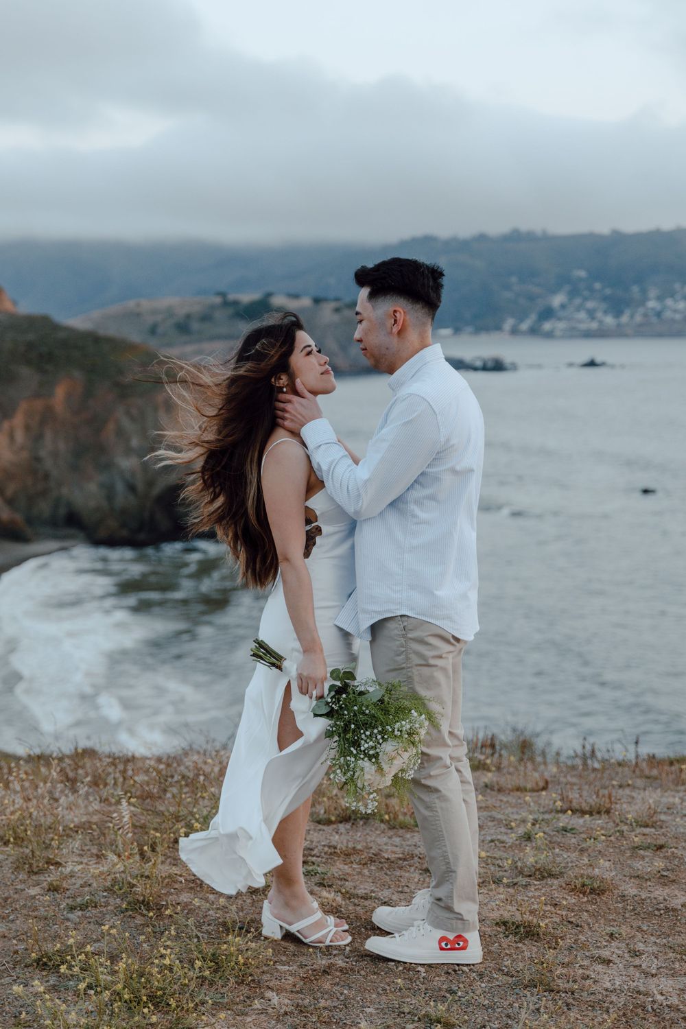 Romantic couple in white embraces on coastal cliff during moody sunset overlooking ocean and mountains.