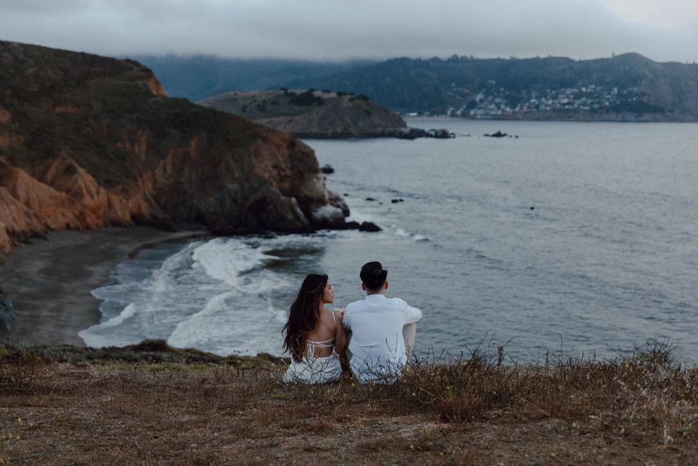 Two people sit on a cliff overlooking dramatic coastal views with crashing waves and rugged shoreline at dusk.