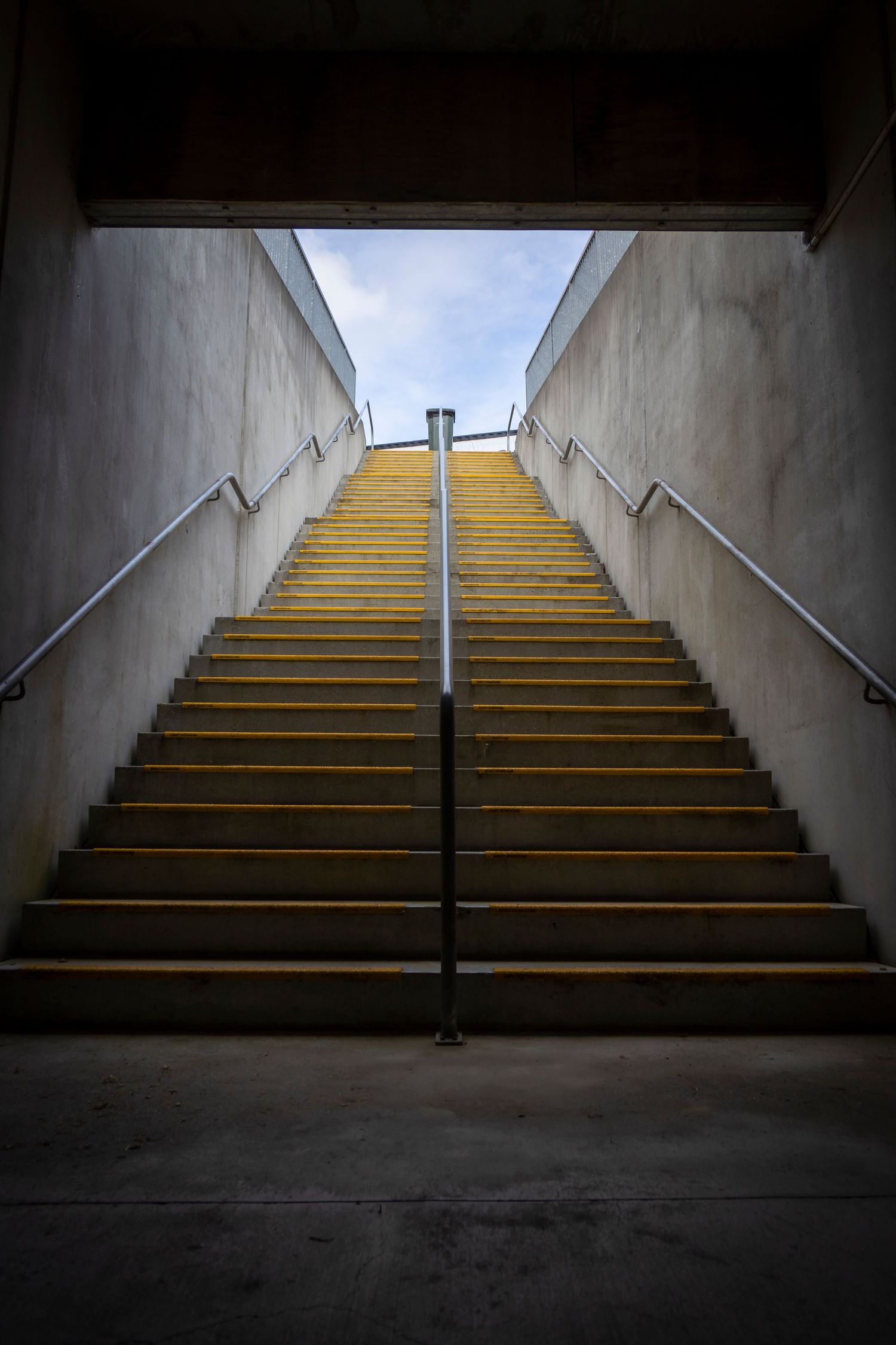 Concrete stairwell with yellow steps leading upward toward daylight, framed by walls and a central handrail.