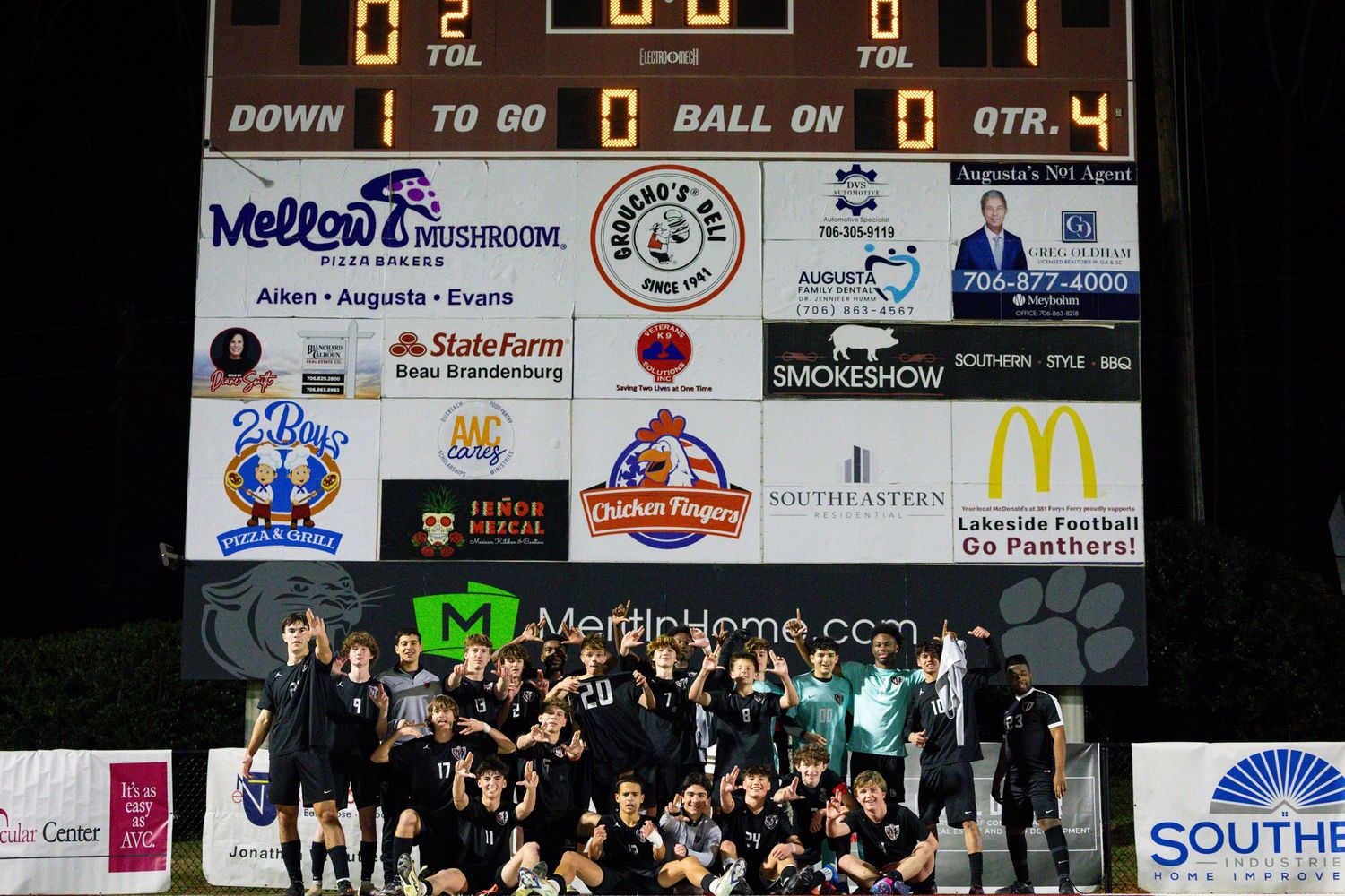 Sports team poses together for group photo on field at night with scoreboard visible.