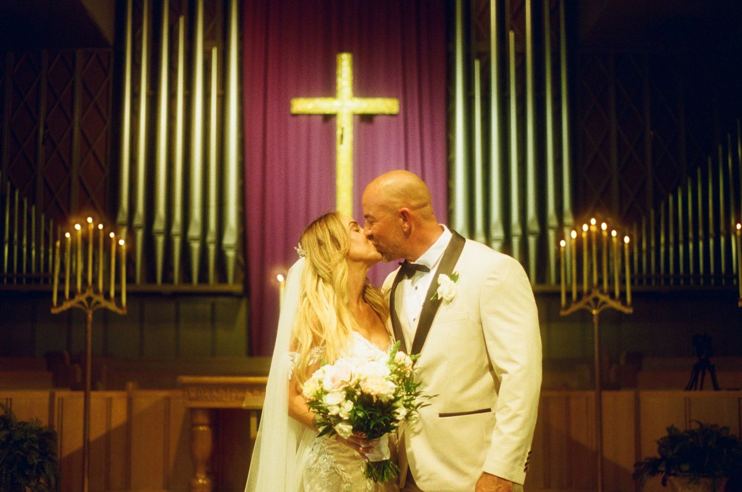 A couple shares a romantic kiss at the altar during their wedding ceremony with organ pipes and cross in the background.