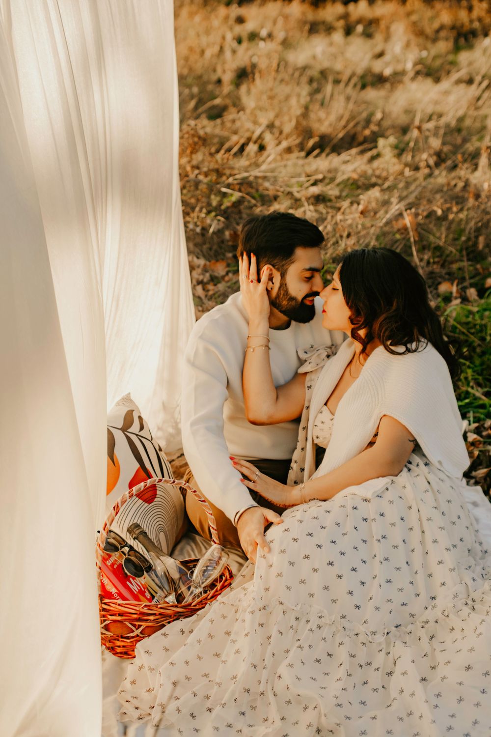 A romantic picnic scene at sunset with a couple sharing an intimate moment beside a white backdrop and picnic basket.