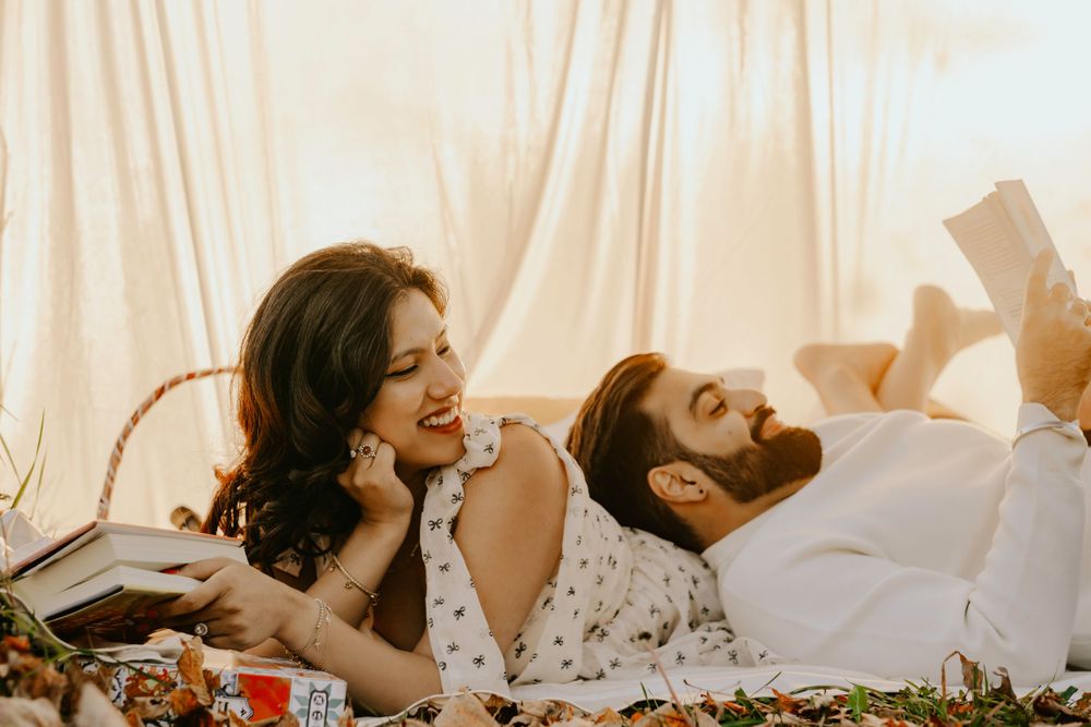 Two people laying on a bed with flowers, laughing and reading books together in warm lighting.