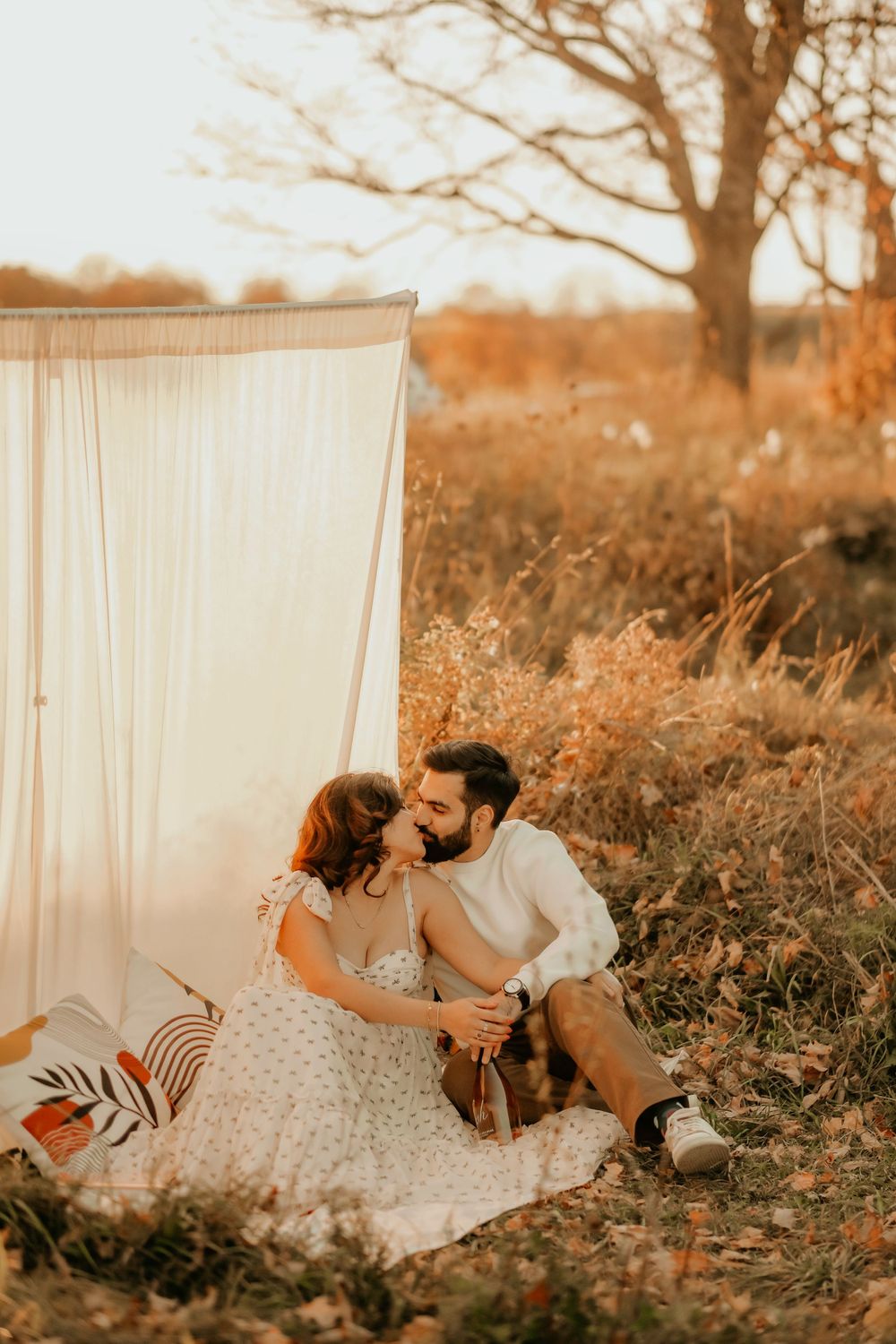 A couple shares an intimate moment on a blanket in a golden field during sunset with a white backdrop behind them.