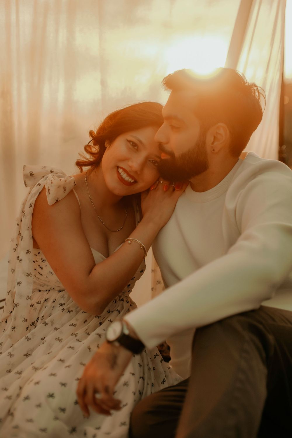 A couple shares an intimate moment in warm lighting as they sit together, with her wearing a polka dot dress.