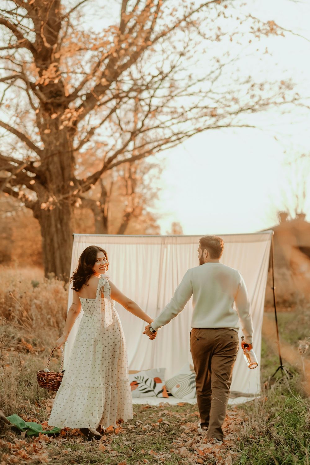 A couple holds hands while walking near a white backdrop in an autumn field with warm golden lighting.