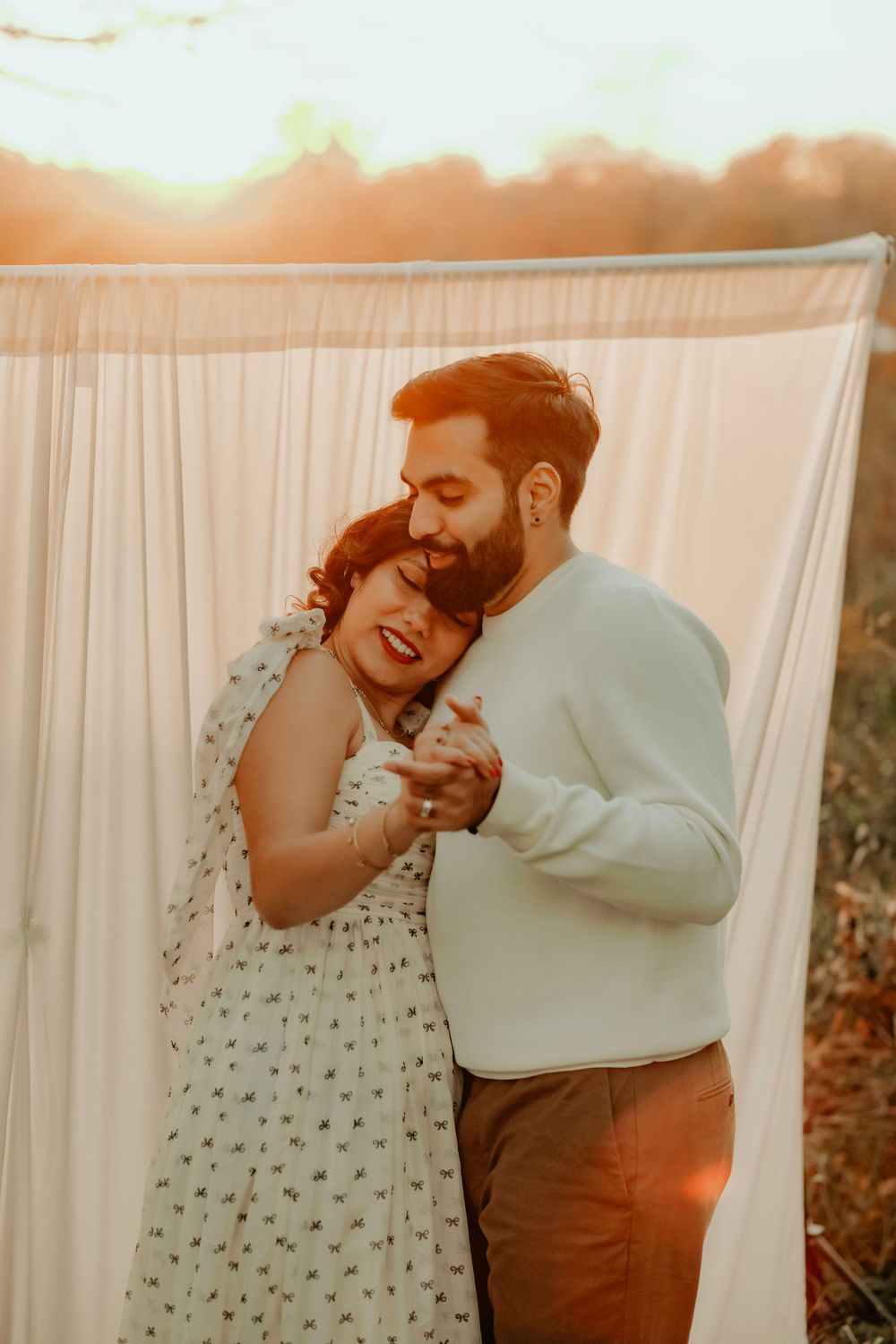 A couple embraces intimately in front of a white curtain backdrop during sunset with warm golden lighting.