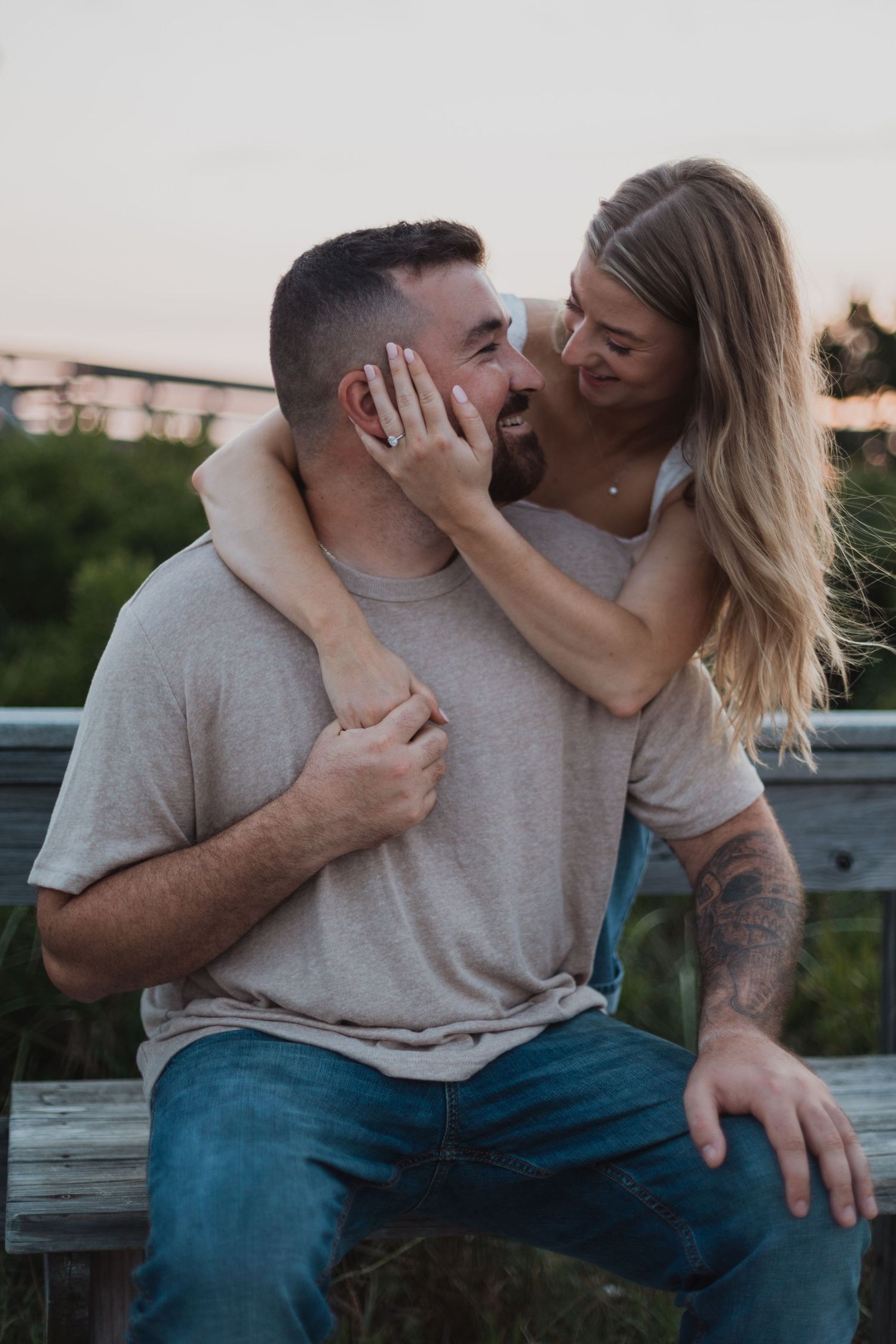 A romantic couple embraces on a sandy beach during sunset, with beachgrass and dunes in the background.