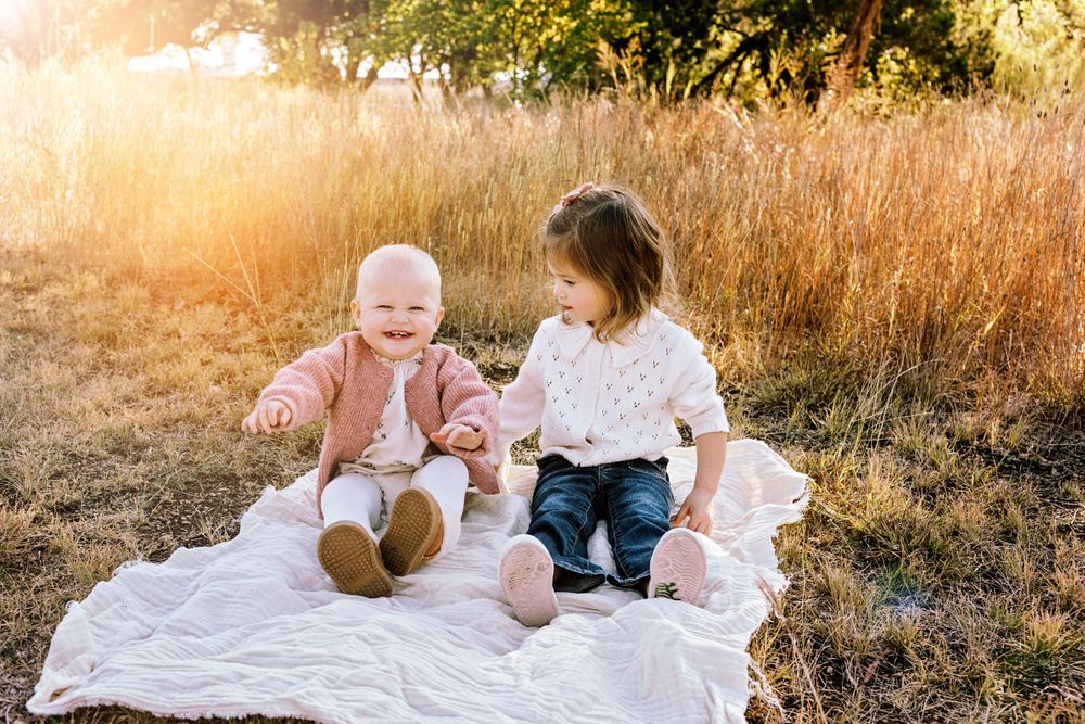 Two children sit together on a white blanket in a golden wheat field during sunset.