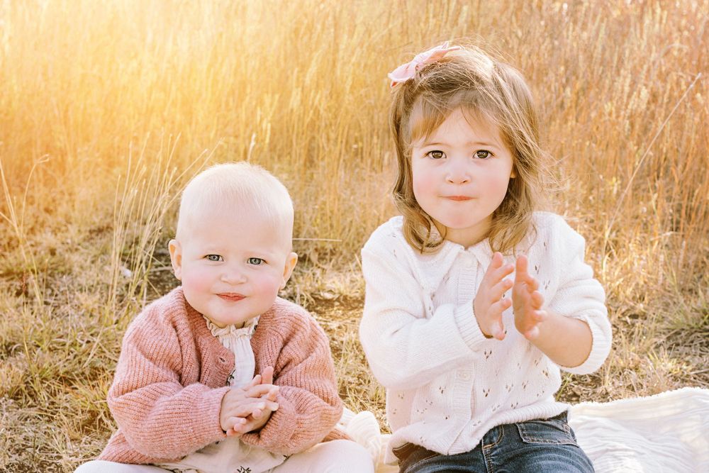 Two children sit together outdoors in a sunlit field with golden grass in the background during sunset.