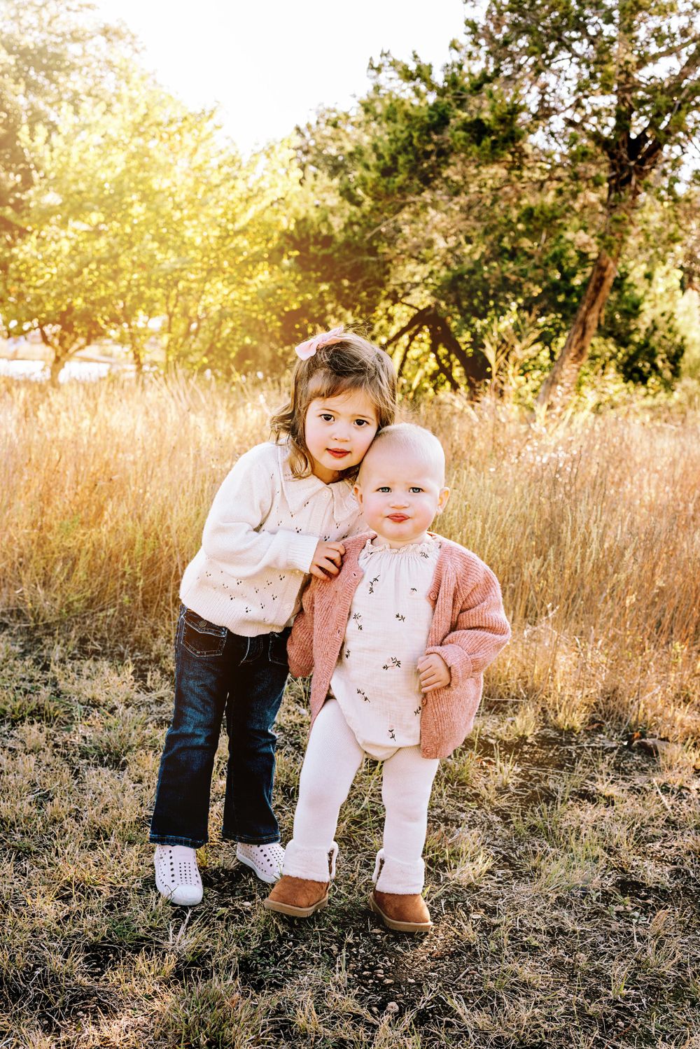Two young children standing together in a golden sunset field with tall grass and trees in the background.