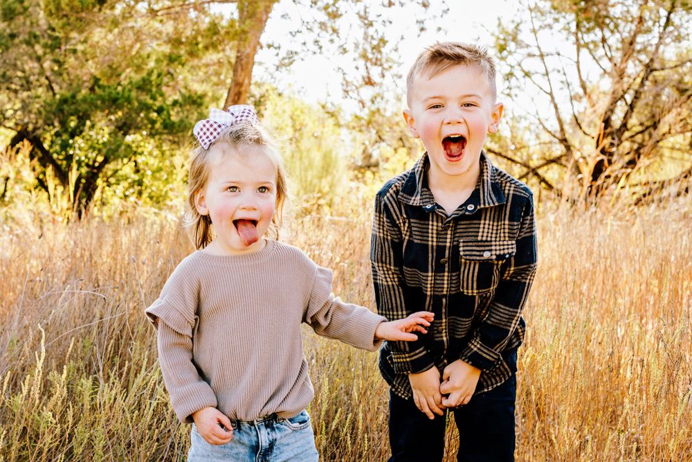 Two young children laugh joyfully while standing together in a golden sunlit field of tall grass.