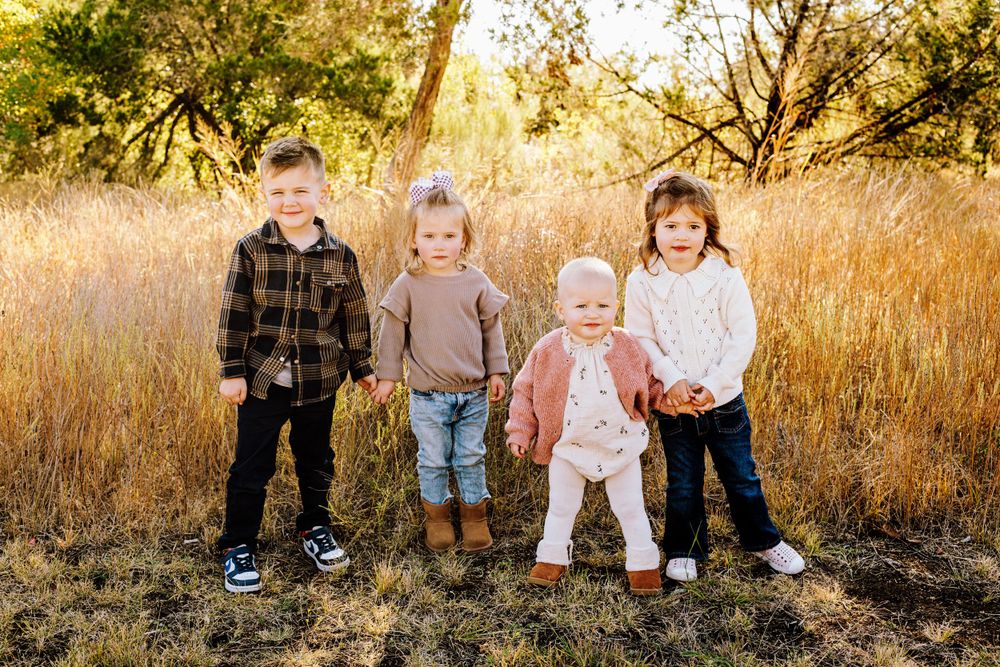 Four young children standing together and smiling in a sun-lit field with dry grass and trees behind them.