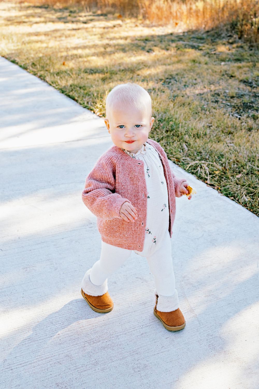 Small child wearing a pink knit cardigan and white pants stands on a concrete path in autumn sunlight.