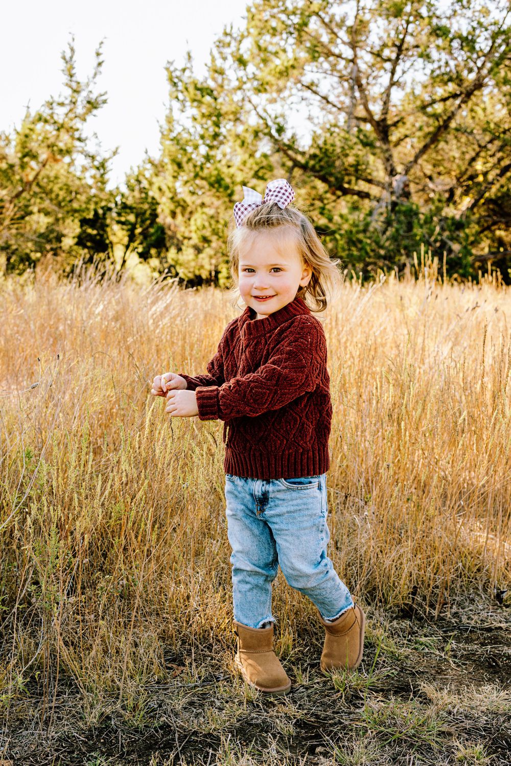 Small child wearing maroon sweater and jeans stands in golden autumn field with evergreen trees in background.