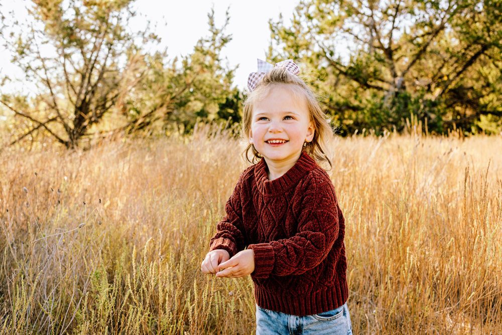 A joyful child wearing a burgundy sweater stands in a golden field of tall grass during sunset.