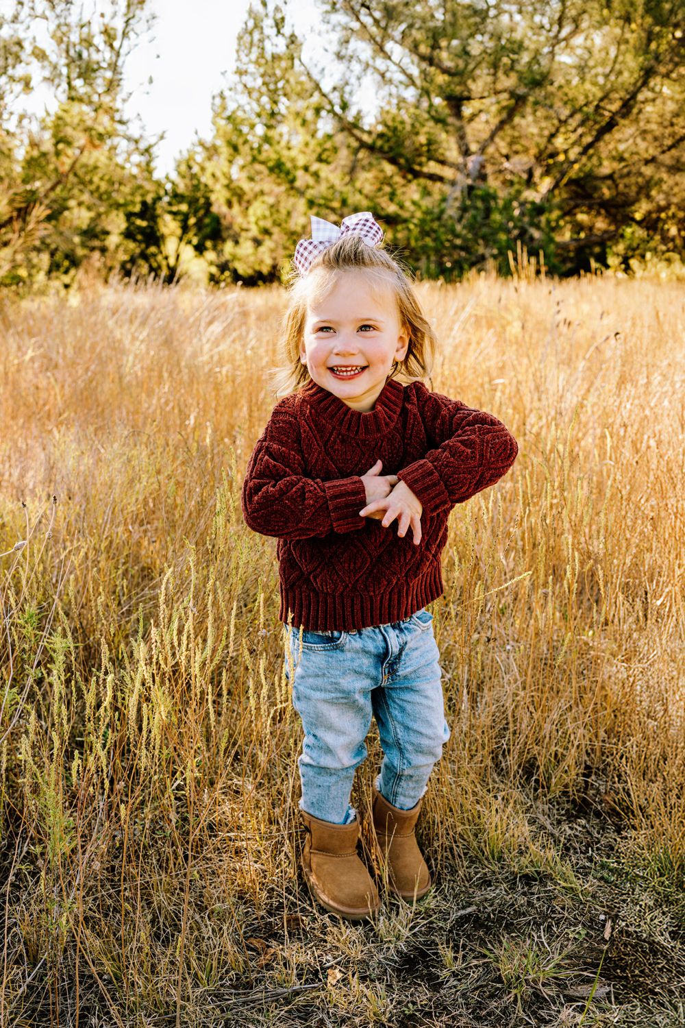 Small child wearing maroon sweater and jeans stands joyfully in golden wheat field during sunset.