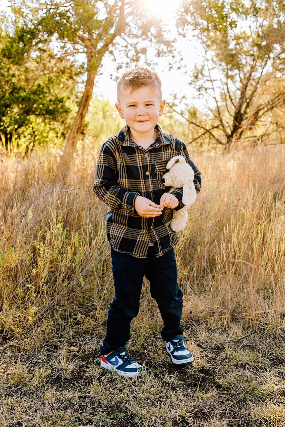 Child in plaid shirt and blue sneakers stands in tall grass holding stuffed toy during golden hour.