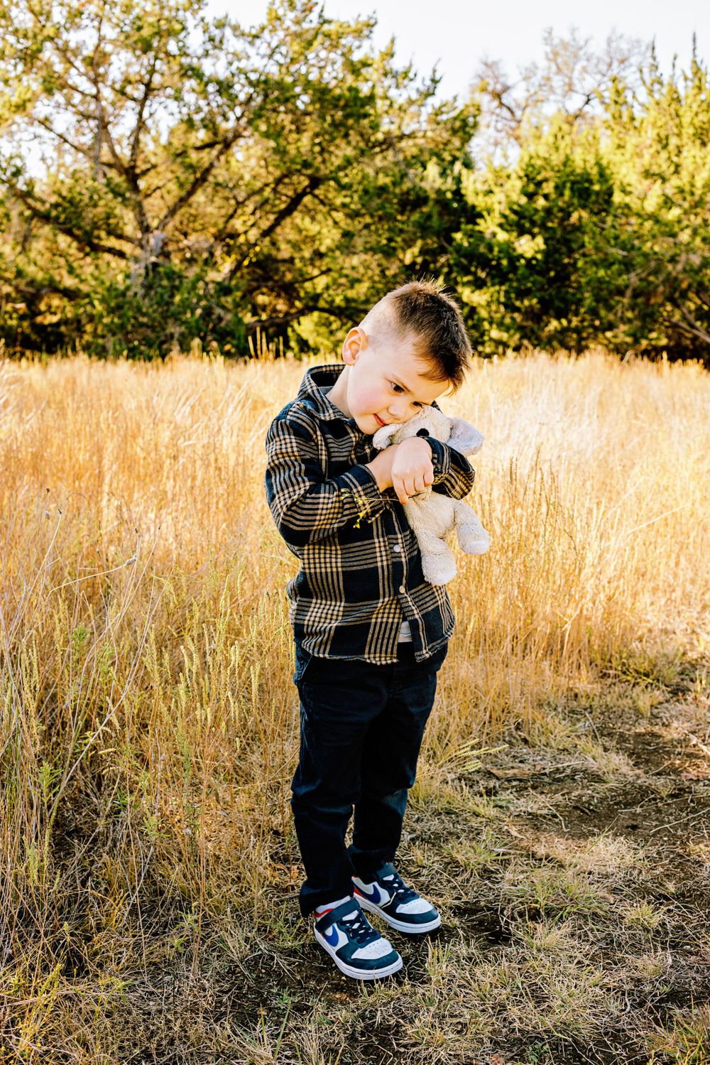 A young child stands in a golden autumn field wearing a plaid flannel shirt, dark pants and blue shoes.